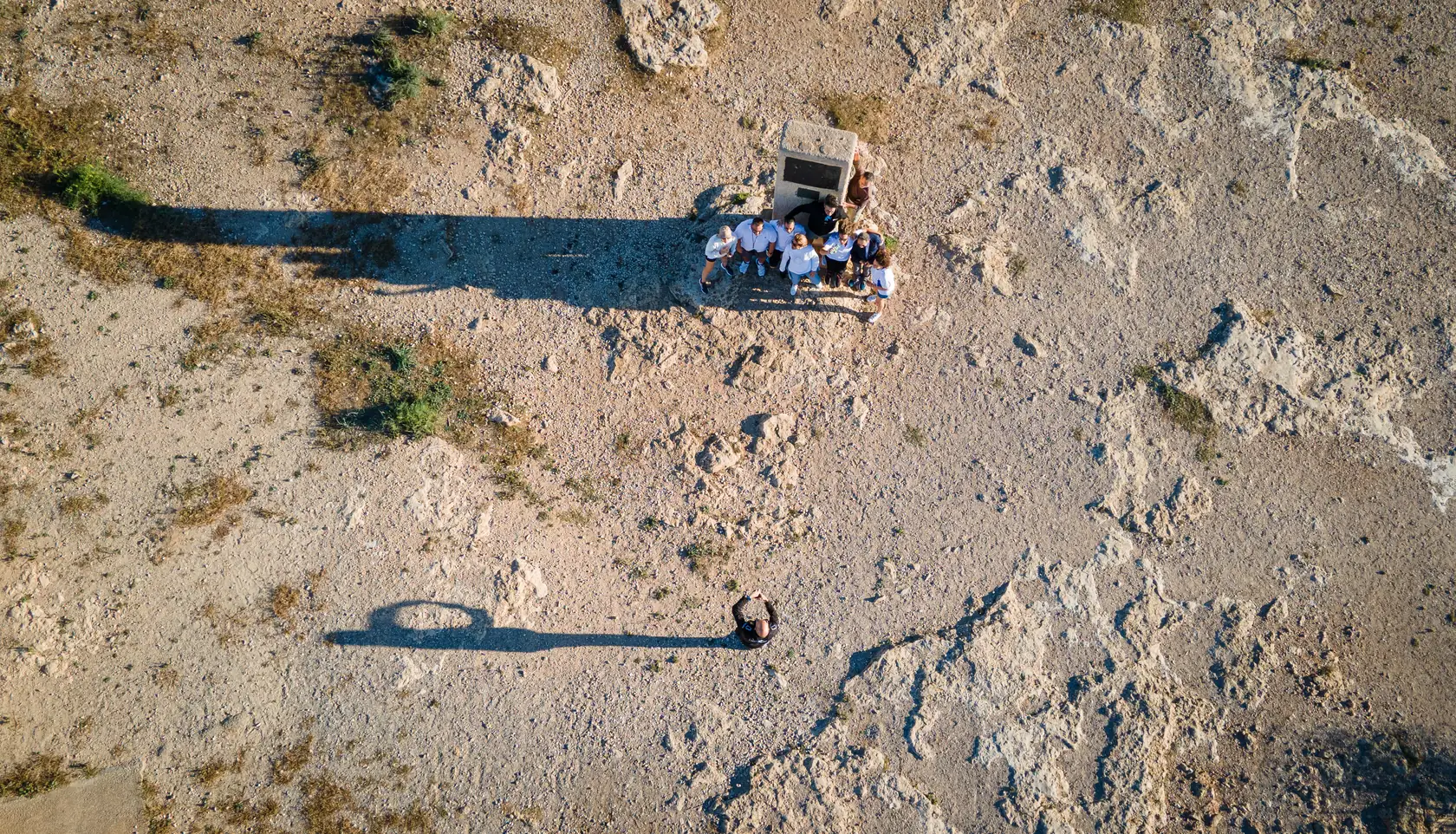 Vista aérea cenital de un pequeño grupo de participantes reunido junto a una caseta de piedra sobre el terreno rocoso.