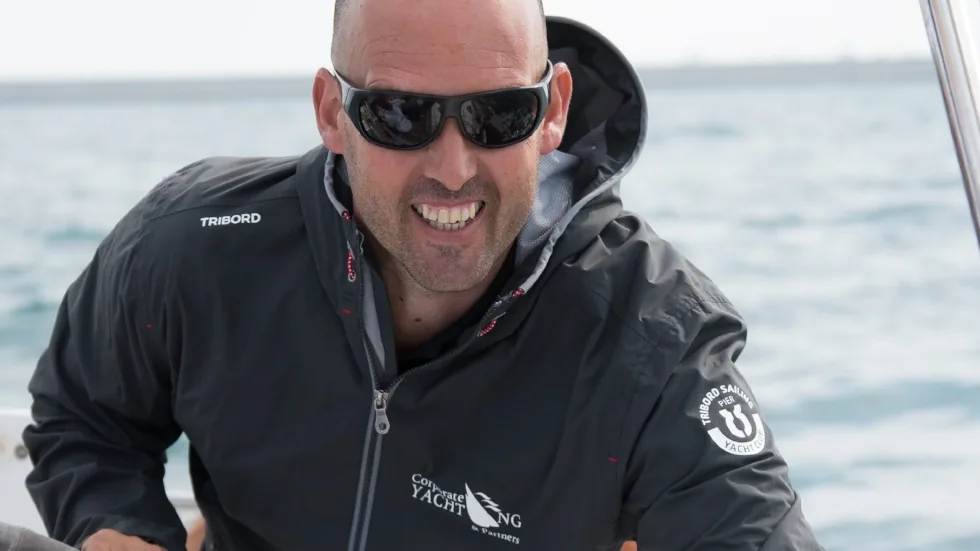 Tripulante sonriente con chaqueta técnica de navegación, con el mar al fondo