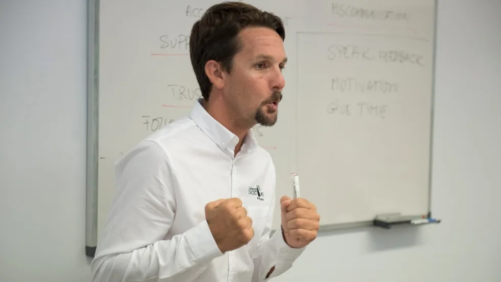 Formador gesticulando con los puños ante una pizarra con conceptos de comunicación y motivación de equipos