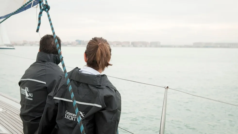 Dos participantes con chaqueta de Corporate Yachting contemplando la costa desde la cubierta del velero