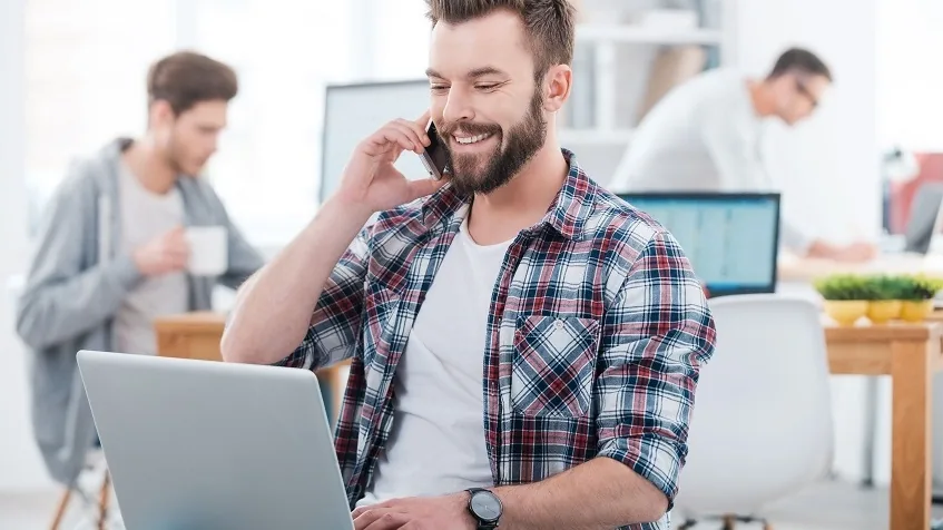 Profesional sonriente hablando por teléfono junto a su portátil, con compañeros trabajando al fondo