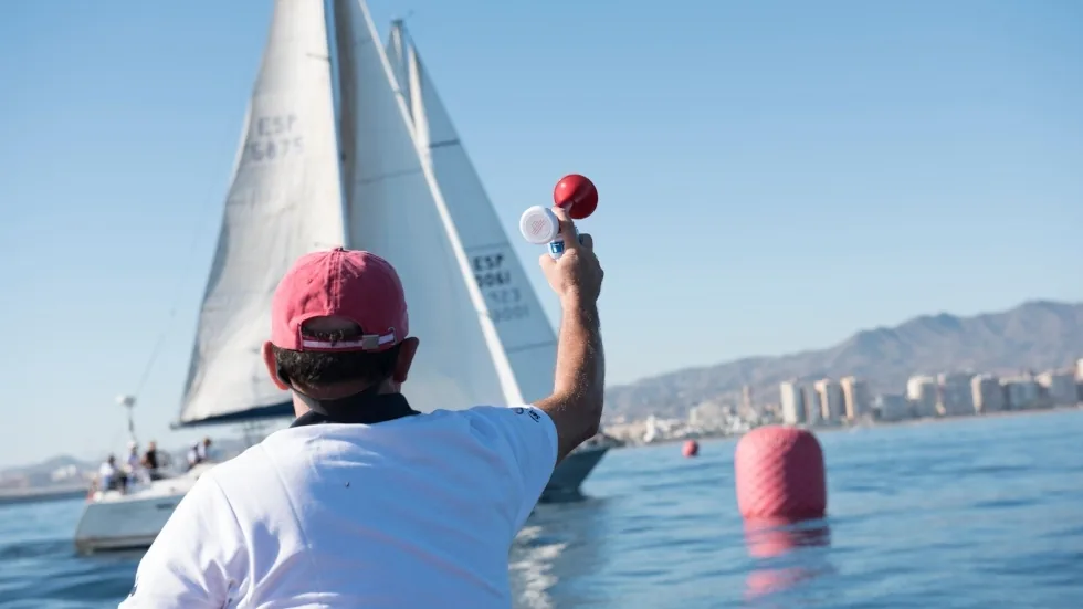 Oficial de regata con gorra roja controlando el crono de salida junto a las boyas