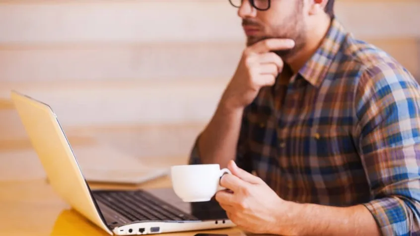 Profesional pensativo con la mano en la barbilla frente al ordenador portátil, taza de café en mano